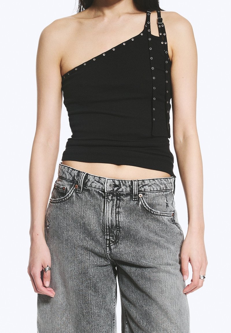 Femme portant un top noir à une épaule avec des œillets métalliques et un jean gris taille haute, montrant un ventre avec des bagues aux doigts.