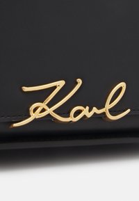 Sac en cuir noir avec une signature dorée en relief "Karl", présentant des lignes épurées et une texture lisse. Accentué par des coutures fines.