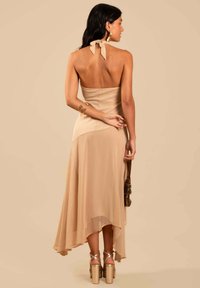 Beige halterneck jurk met een asymmetrische zoom, zachte stof en een gelaagd ontwerp. Model draagt zilveren sleehakken en een kleine tas.