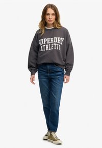 Szary sweatshirt z napisem "SUPERDRY ATHLETIC", zestawiony z niebieskimi dżinsami i oliwkowymi trampkami. Luźny krój i casualowy design.