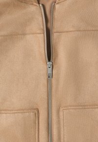 Veste en daim beige avec fermeture éclair en métal argenté partiellement fermée et deux poches carrées à l'avant avec des coutures visibles.