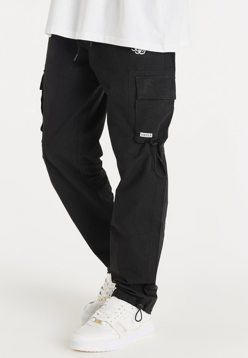 Pantalon cargo noir avec poches latérales et cordons ajustables aux chevilles, porté avec des baskets blanches et un haut blanc.