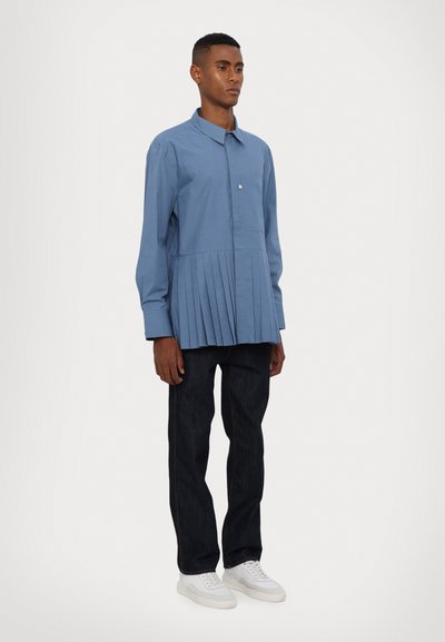 Chemise bleu à manches longues avec un devant plissé, fermeture à boutons et une poche sur la poitrine, associée à un jean noir et des baskets blanches.