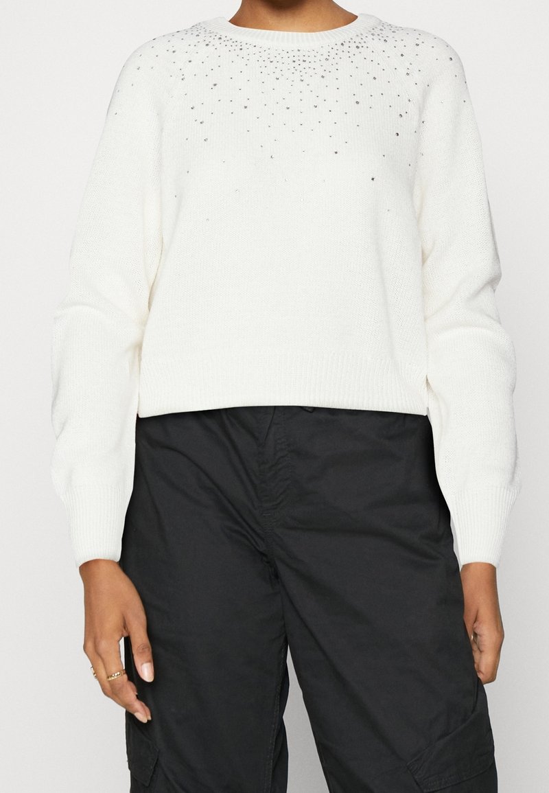 Pull en maille blanc avec de petites ornements argentés dispersés sur le haut devant, porté avec un pantalon style cargo noir.
