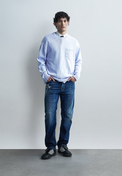 Jeune homme debout, les mains dans les poches, portant un pull bleu clair par-dessus une chemise blanche, un jean bleu déchiré et des baskets noires à rayures blanches.