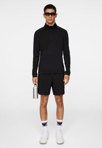 Svart atletisk zip-up-tröja med en framficka, tillsammans med svarta shorts. Modellen håller en silverfärgad vattenflaska, bär solglasögon och vita sneakers.