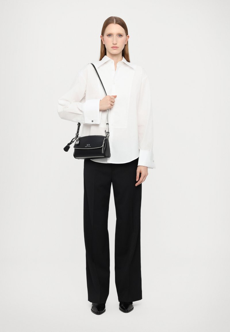 Camicia bianca con colletto oversize, pantaloni neri a gamba larga e una piccola borsa nera con accenti argentati, su uno sfondo neutro.