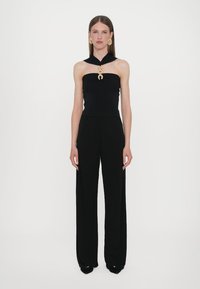 Schwarzer ärmelloser Jumpsuit mit Halterneck und goldener Hummerverschlussdetails, kombiniert mit großen goldenen Ohrringen, hat eine lockere Passform und weite Beine.