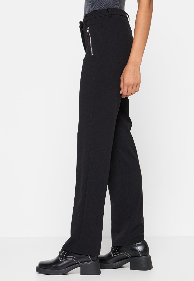 Pantalons noirs à jambes larges en tissu lisse. Dotés d'une poche zippée latérale élégante et d'une taille ajustée. Associés à des chaussures noires.