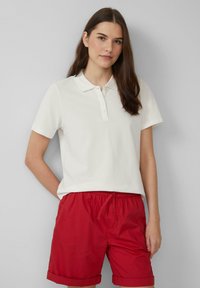 Witte poloschort met korte mouwen en een kraag, voorzien van een knopenlijst. Gecombineerd met rode shorts met opgerolde zoom. Glad stof.