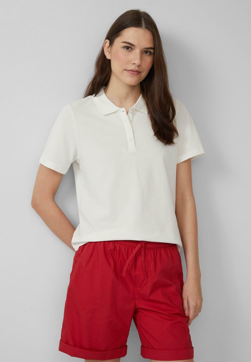 Witte poloschort met korte mouwen en een kraag, voorzien van een knopenlijst. Gecombineerd met rode shorts met opgerolde zoom. Glad stof.
