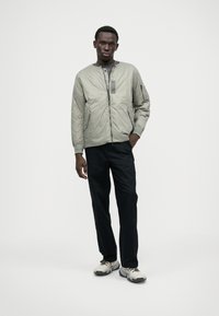 Chaqueta bomber gris claro con cremallera, cuello acanalado y bolsillos laterales. Pantalones negros y zapatillas grises complementan el conjunto.