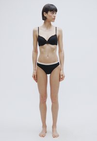 Μαύρο δαντελένιο σουτιέν με ρυθμιζόμενους ιμάντες και διακοσμητικό φιόγκο, σε συνδυασμό με μαύρο σλιπ με αντίθετη λευκή μέση, φορεμένα από μια γυναίκα.