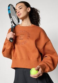 Narancssárga cropped pulcsi, amelyen meg van emelve a "NikeCourt" logó, fekete szoknyával párosítva. Zöld teniszlabdát és fekete teniszütőt tart.