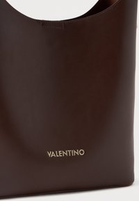 Brązowa skórzana torba typu tote o gładkiej fakturze z złotym logo "Valentino" z przodu. Posiada zakrzywiony kształt u góry.