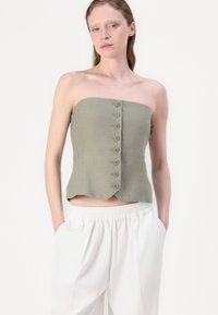 Oliven grøn strapless topp med en tettsittende design, åtte knappedetaljer nedover fronten, og en bølget hemkant, sammen med hvite bukser.