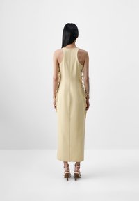 Femme aux longs cheveux foncés portant une robe midi beige sans manches avec des liens sur les côtés et des sandales à talons hauts lacées, debout face à un mur blanc.
