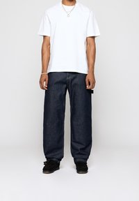 Valkoinen puuvillapaita, löysät tummansiniset farkut ja mustat kengät. Huomionarvoisia yksityiskohtia ovat hopeaketju ja näkyvät ompeleet.