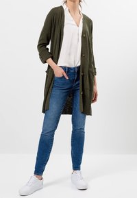 Olivově zelený pletený kardigan se třemi knoflíky, nošený rozepnutý přes bílý top. Kombinován s úzkými modrými džíny a bílými teniskami.