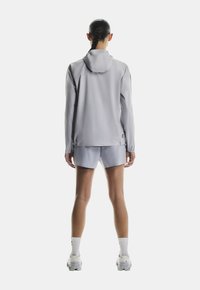 Siva športna jakna z kapuco, v kombinaciji s svetlobno sivimi kratkimi hlačami. Jakna ima gladko teksturo in podrobnost logotipa na hrbtni strani.
