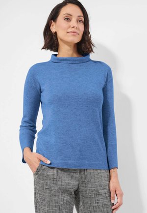 Femme aux cheveux foncés mi-longs portant un pull bleu à manches longues et un pantalon gris à carreaux, une main dans la poche, debout devant un fond blanc.