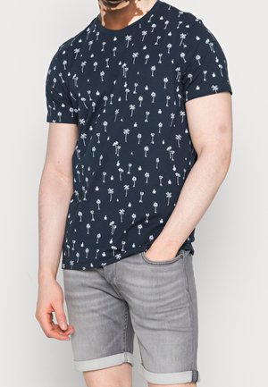 Homme portant un t-shirt bleu marine avec des imprimés de palmiers blancs et un short en jean gris retroussé, une main dans la poche, sur fond uni.