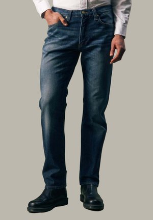 Mann trägt dunkelblaue Straight-Fit-Jeans, schwarze Lederstiefel und ein weißes Langarmhemd mit einer Hand in der Tasche.