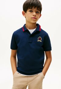 Marineblå polo T-shirt med røde krave accenter, prydet med et broderet våbenskjold og korte ærmer, parret med beige shorts.