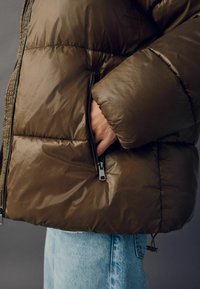 Brun pufferjakke med en glansfuld finish, der har lynlåslommer og quiltede sektioner. Båret over lyseblå denimjeans.