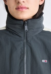 Chaqueta de cremallera gris oscuro con cuello alto, que presenta un panel en el hombro en blanco contrastante y un pequeño logo bordado en el pecho. Textura suave.