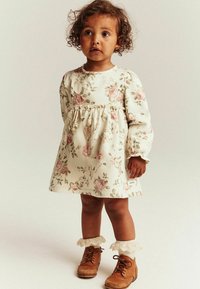 Kukka-aiheinen mekko pitkillä hihoilla, creme-harmaalla taustalla vaaleanpunaisia ruusuja. Koristeltu pitsireunaisilla sukilla ja ruskeilla nauhakengillä.