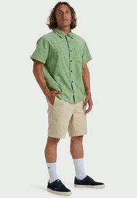 Camicia a maniche corte, di colore verde chiaro, con bottone e texture a motivi, abbinata a pantaloni corti beige, calze bianche e sneakers blu navy.