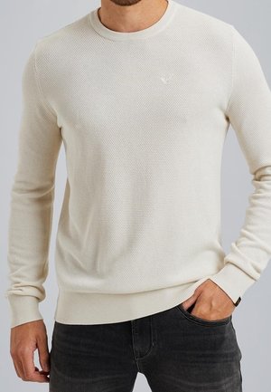 Mężczyzna ubrany w kremowy, teksturowany sweter dzianinowy z prążkowanymi mankietami i dołem, w połączeniu z ciemnoszarymi dżinsami, z ręką w kieszeni.