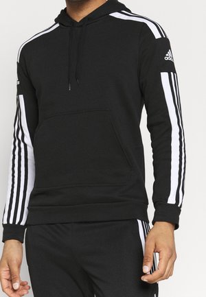 Mężczyzna ubrany w czarną bluzę z kapturem Adidas z białymi paskami na rękawach i dopasowane czarne spodnie z białymi paskami po bokach, stojący na jednolitym tle.