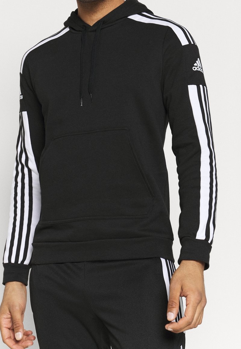 Homme portant un sweat à capuche Adidas noir avec des bandes blanches sur les manches et un pantalon noir assorti avec des bandes blanches latérales, debout devant un fond uni.