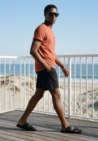 Homme portant des lunettes de soleil, un t-shirt orange, un short noir et des sandales marchant sur une terrasse en bois près d'une plage avec l'océan et des dunes de sable en arrière-plan.