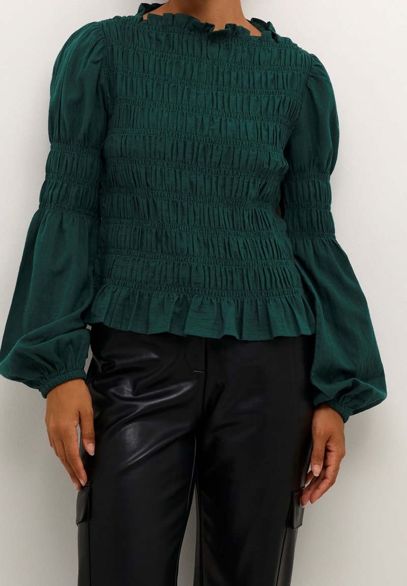 Femme portant un chemisier smocké vert foncé avec de longues manches bouffantes et un pantalon en cuir noir, debout devant un fond blanc uni.