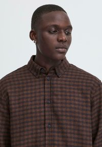 Camisa de cuadros marrón con collar abotonado, botones negros y una tela texturizada. El patrón presenta un diseño de cuadrícula en varios tonos.