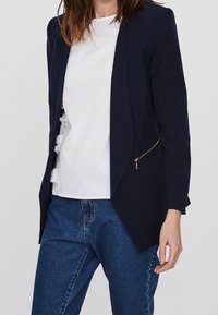 Námořnický blazer s upnutým střihem, strukturovaným tvarem, jednou zipovou kapsou a hladkým materiálem, vrstvený přes bílou volánkovou blůzu a džíny.