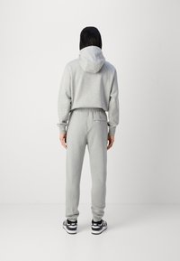 Osoba oblečená v světle šedé mikině s kapucí a odpovídajících teplácích, stojící zády, s černobílými teniskami Nike.