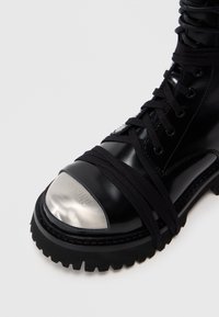 Čierna kožená topánka s kovovou špičkou, hrubou gumovou podrážkou a čiernymi šnúrkami omotanými okolo členku, s textúrovaným povrchom.