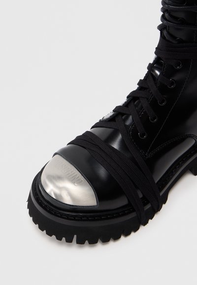 Stivale in pelle nera con punta metallica, suola in gomma spessa e lacci neri avvolti attorno alla caviglia, caratterizzato da dettagli di superficie texturizzati.
