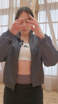 Femme en tenue de sport avec une veste grise et un crop top blanc ajuste ses cheveux dans une pièce avec des rideaux transparents et un sol en bois.