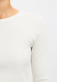 Fehér bordázott hosszú ujjú felső kerek nyakkivágással, testhezálló szabással, lágy tapintású anyaggal és finom, függőleges csíkos mintával.