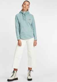 Világoskék kapucnis pulóver texturált mintával, elülső zsebbe és állítható kapucnival, fehér, bő szárú nadrággal és fekete fűzős csizmával párosítva.