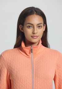 Perzikkleurige zip-up jack met een gestructureerd patroon, staande kraag en zilveren rits. De stof lijkt glad met subtiele details op het oppervlak.
