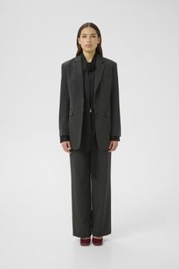 Vrouw staat naar voren gekeerd in een donkergrijze oversized blazer en broek, zwarte blouse met bloemmotief op de hals, en bordeauxrode schoenen.