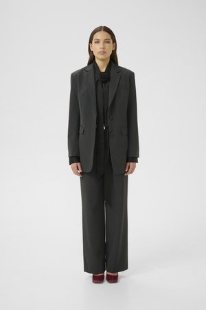 Kvinne står vendt mot kameraet iført en mørkegrå oversized blazer og bukser, svart bluse med blomsterdetaljer ved halsen, og burgunder sko.