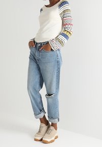 Valkoiset ribbattu pitkähihainen paita, jossa on moniväriset kuviolliset hihat, yhdistettynä vaaleansinisiin kulutettuihin farkkuihin ja beigekirjavien platósneakereiden kanssa.