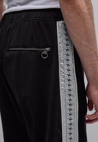 Μαύρο παντελόνι με ελαστική μέση, πίσω τσέπη με φερμουάρ και κρίκο, καθώς και αντανακλαστική ασημί λωρίδα στο πλάι με επαναλαμβανόμενες μαύρες σιλουέτες πουλιών.